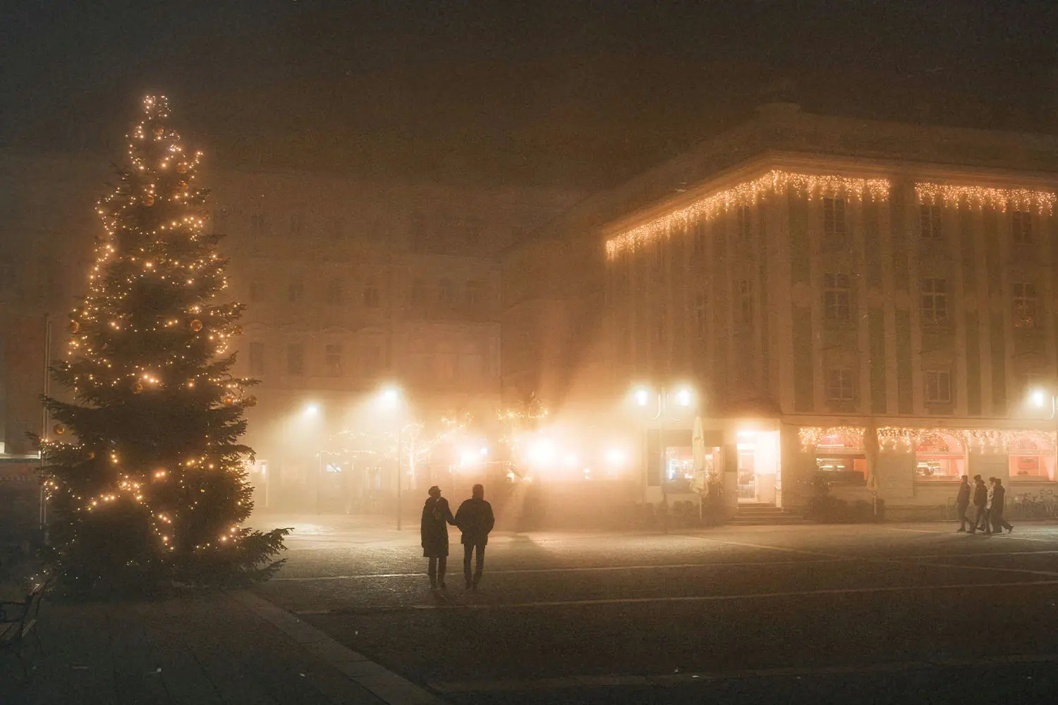 two people walking down lit street with snow all around and a lit christmas tree