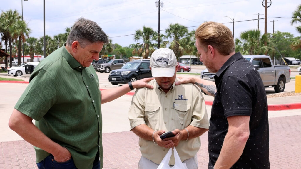 On a sunny day, two men are laying their hands on another man's shoulders, praying for him, while standing in a Kerrville parking lot.