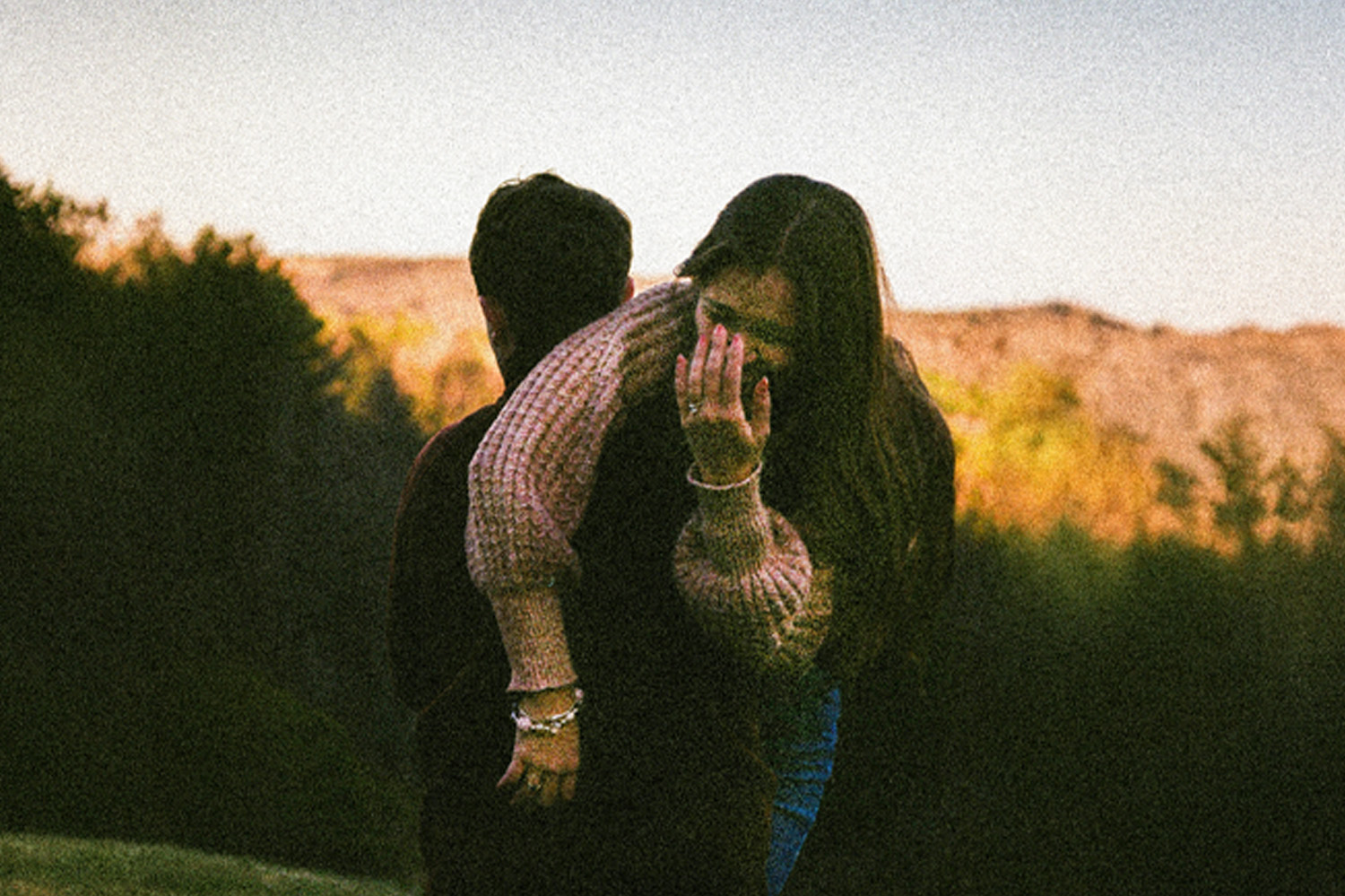 Two people outdoors, one carrying the other on their back in a grassy area with trees and hills under a clear sky.