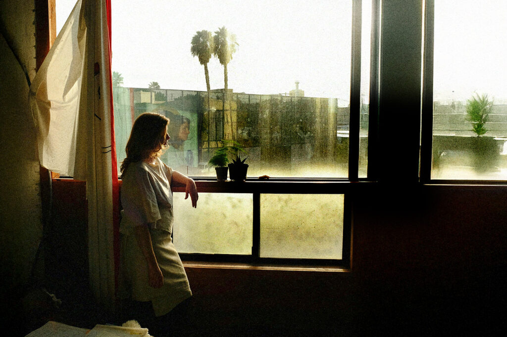 BelieveInMiracles-Blog-KCM - KCM Blog Person standing by a sunlit window with plants, gazing outside as Miracles feel possible in a calm urban setting.