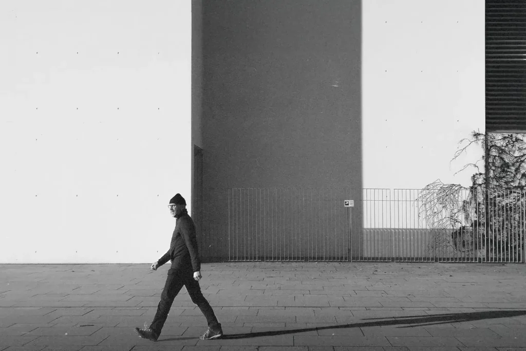 man dressed in black with glasses and a beanie hat walking down the street