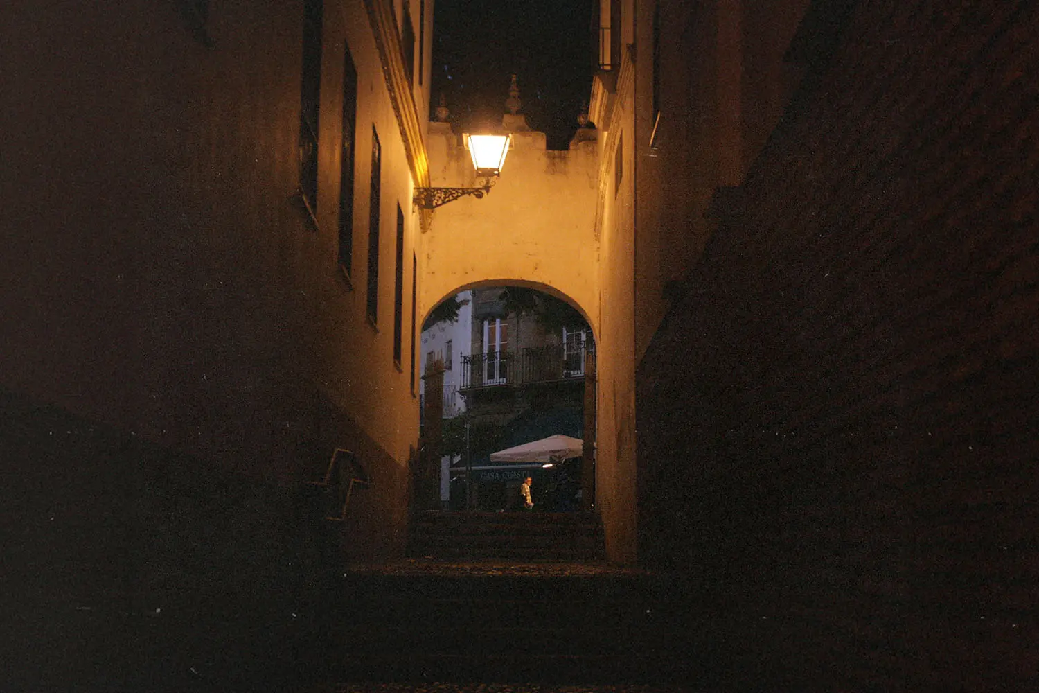 Narrow dimly lit alley leading to an archway with a warm lantern glowing above it at night