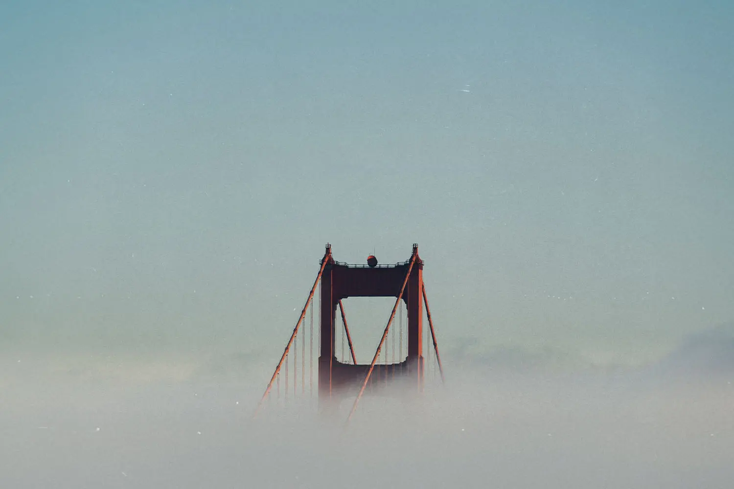 Top of a suspension bridge emerging through thick fog beneath a clear sky