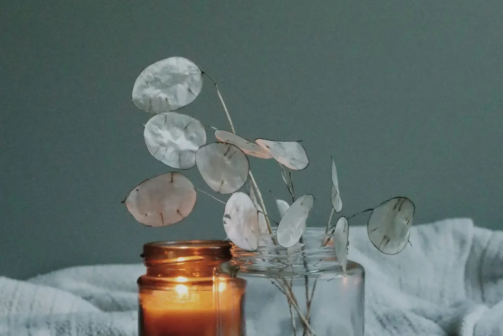 two jars next to each other, one with a lit candle, and one with white silk flower leaves on stems