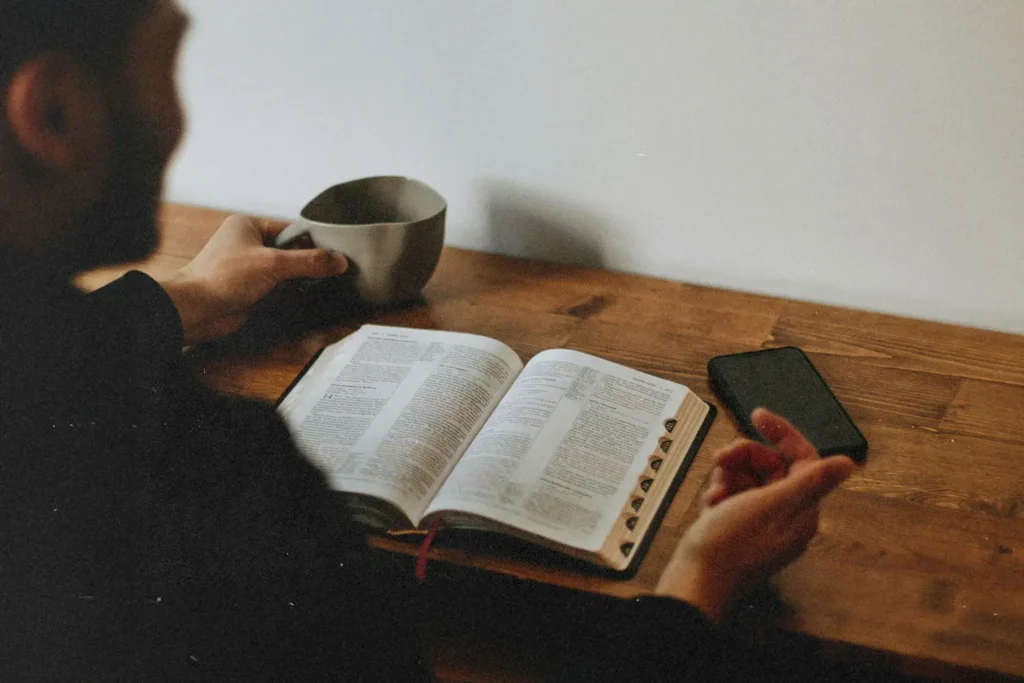 20250107-3-Keys-To-Stop-Living-Paycheck-to-Paycheck-KCM-Blo - KCM Blog Man reading open Bible resting on wooden table with a coffee cup in hand
