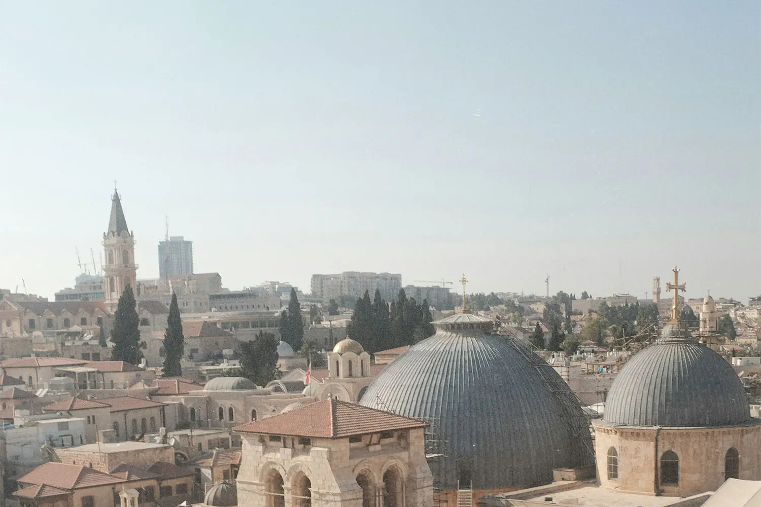 Cityscape featuring domed church roofs, stone buildings, and distant towers under a bright, clear sky