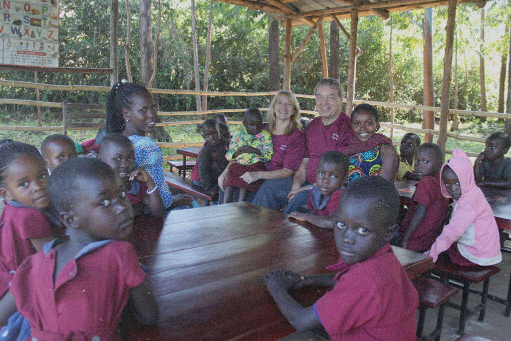 Children gather around missionaries seated at outdoor tables during a Uganda missions class, reflecting their commitment to teaching and discipleship.