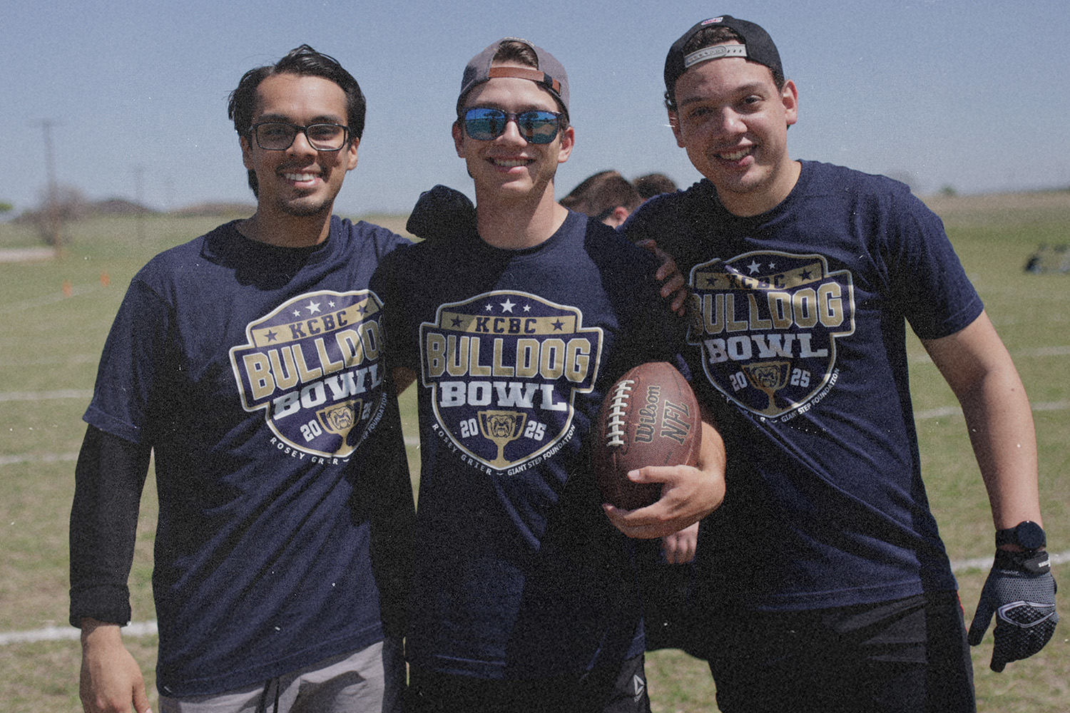 KCBC students stand together on a sunny field wearing Bulldog Bowl shirts, holding a football and reflecting the unity and community found in pursuing God’s plan.