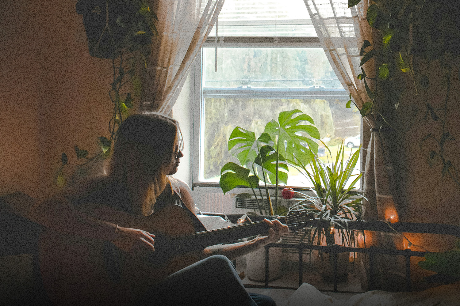 Believer sitting by a sunlit window playing acoustic guitar, surrounded by plants, reflecting consistency over hype and a quiet, steady faith lifestyle.